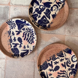 May include: Three ceramic plates with a unique design. The plates feature a cream-colored background with navy blue figures and pink accents. The rims are a speckled, earthy brown. The plates are arranged on a neutral-toned tiled surface.