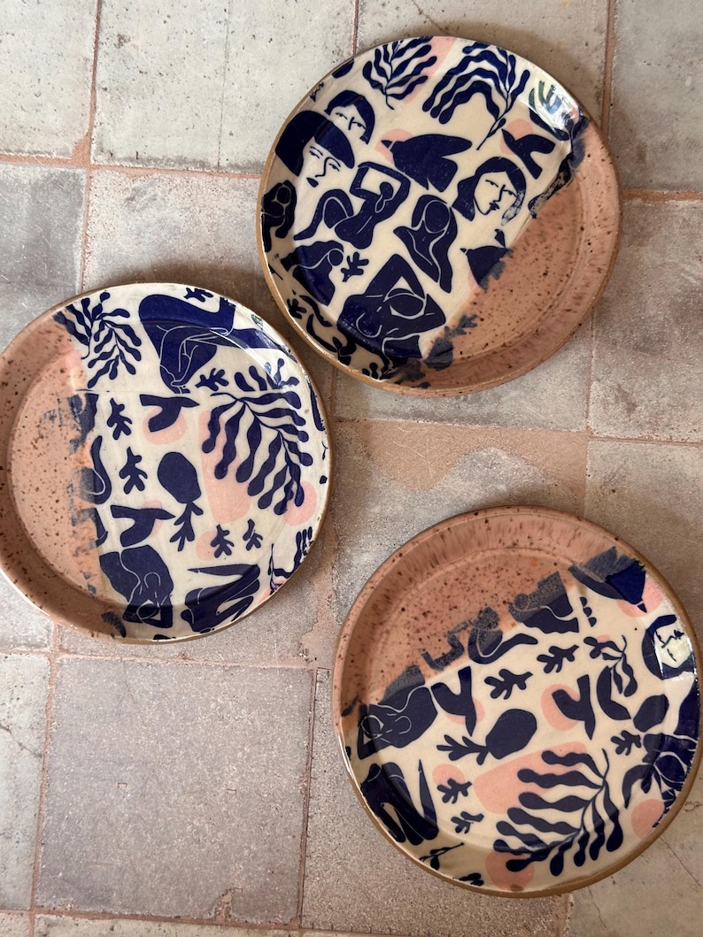 May include: Three ceramic plates with a unique design. The plates feature a cream-colored background with navy blue figures and pink accents. The rims are a speckled, earthy brown. The plates are arranged on a neutral-toned tiled surface.