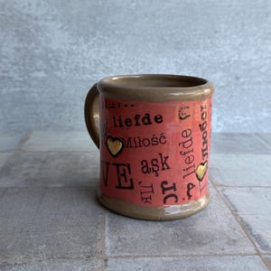 Close-up of coral handcrafted ceramic coffee mug in love language style, showing 'LOVE', 'LIEFDE', 'MILOŚĆ' text and 24k gold hearts on body, perfect as a friendship gift