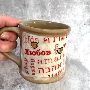 May include: A beige ceramic mug with a brown handle and rim. The mug features the word "Любовь" in red, along with other words in different languages, and gold heart accents. The mug is held by a hand.