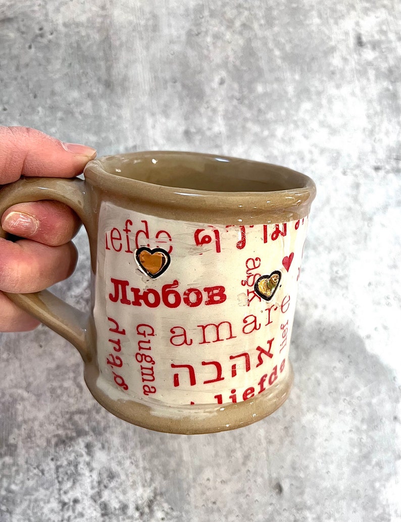 May include: A beige ceramic mug with a brown handle and rim. The mug features the word "Любовь" in red, along with other words in different languages, and gold heart accents. The mug is held by a hand.