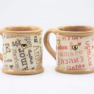 May include: Two ceramic mugs with beige and brown tones. The mugs are decorated with words of love in different languages, gold heart accents, and a beige background. The mugs have a handle.