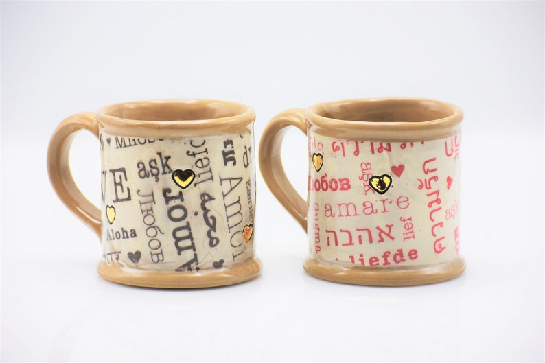 May include: Two ceramic mugs with beige and brown tones. The mugs are decorated with words of love in different languages, gold heart accents, and a beige background. The mugs have a handle.