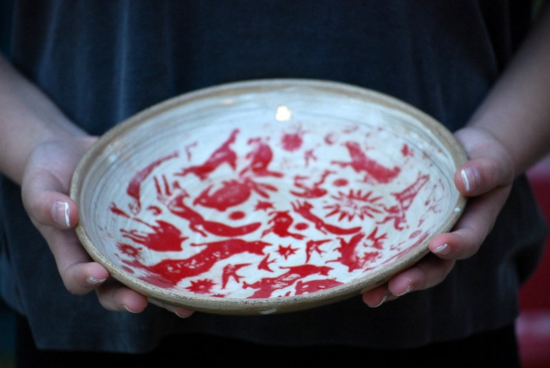 May include: A white ceramic bowl with a red design of stylized animals and geometric shapes.