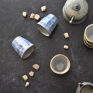 May include: Four blue and white ceramic teacups with gold trim, two are upright and two are upside down.  There are sugar cubes scattered on a dark gray surface.