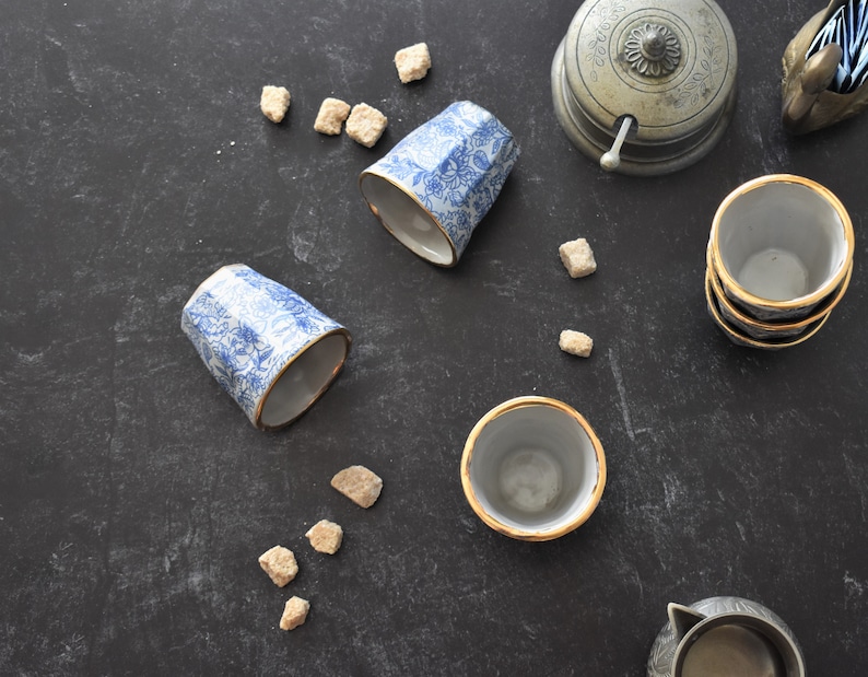 May include: Four blue and white ceramic teacups with gold trim, two are upright and two are upside down.  There are sugar cubes scattered on a dark gray surface.