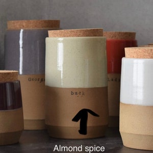 Op de afbeelding: Een verzameling keramische kruidenpotten met kurken deksels. De potten zijn tweekleurig, met een lichtbeige bovenkant en een bruine onderkant. De pot in focus heeft het woord "buck" erop gedrukt, met een pijl omhoog. De tekst "Almond spice" staat onderaan.