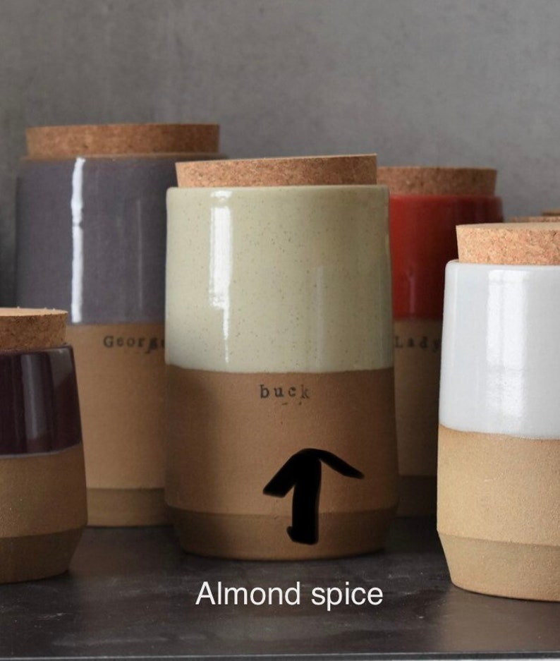Op de afbeelding: Een verzameling keramische kruidenpotten met kurken deksels. De potten zijn tweekleurig, met een lichtbeige bovenkant en een bruine onderkant. De pot in focus heeft het woord "buck" erop gedrukt, met een pijl omhoog. De tekst "Almond spice" staat onderaan.