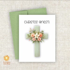 May include: A white card with green trim and the text "Christos Anesti" in black. A white cross with a light green background is decorated with pink and white flowers.