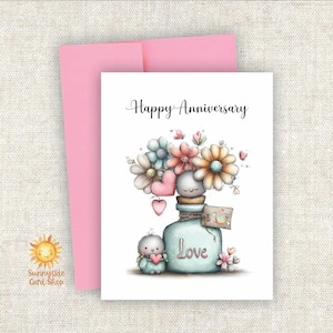 May include: A greeting card with the words "Happy Anniversary" in black script. The card features a whimsical illustration of a bottle filled with flowers and two bird-like figures. A pink envelope is behind the card.