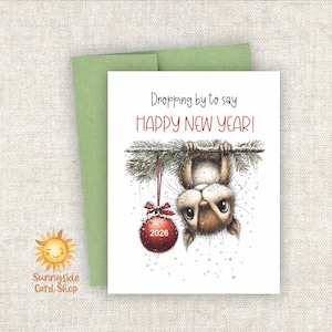 May include: A white greeting card with a watercolor illustration of a squirrel hanging upside down from a branch, with a red ornament that says "2026". The text on the card reads "Dropping by to say HAPPY NEW YEAR!" A green envelope is behind the card.