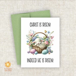 May include: A white card with a green envelope. The card has a white lamb sleeping in a basket of flowers. The text on the card reads "Christ is Risen!" and "Indeed He is Risen!"