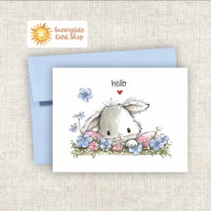 May include: A greeting card with a watercolor illustration of a fluffy white bunny peeking out from behind blue and pink flowers. The word "hello" is written above the bunny, with a red heart. A light blue envelope is behind the card.