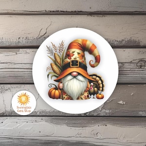 May include: A round sticker featuring a Thanksgiving gnome with a long white beard, wearing an orange and brown hat and holding a pumpkin. A turkey, corn, and pumpkins are also featured. The sticker has the text "Sunnyside Card Shop".