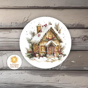 May include: A round sticker featuring a watercolor illustration of a gingerbread house covered in snow. The house has a red and white striped candy cane door, holly accents, and a snowy roof. The sticker also has the text "Sunnyside Card Shop".