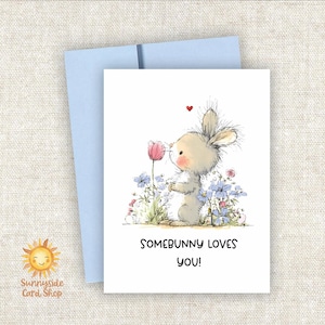 May include: A greeting card with a light blue envelope. The card features a cute bunny smelling a pink tulip, surrounded by blue and pink flowers. Text on the card reads "SOMEBUNNY LOVES YOU!" and has a small red heart above the bunny.