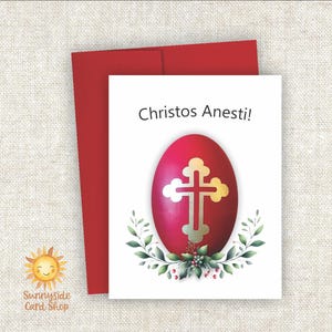 May include: A red Easter card with a matching red envelope. The card features a red egg with a gold cross and the words "Christos Anesti!" The egg is surrounded by green leaves and berries. The SunnySide Card Shop logo is in the lower left corner.
