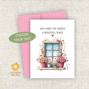 May include: A greeting card with a pink envelope featuring a wooden window illustration surrounded by colorful potted flowers and the text, YOU MAKE THE WORLD A BEAUTIFUL PLACE, with a small red heart above the window frame and a green oval label.