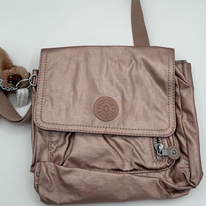 May include: A metallic rose-gold shoulder bag with a flap closure and a small front zippered pocket. The bag features a circular logo patch and a matching shoulder strap. A small monkey keychain hangs from the side.