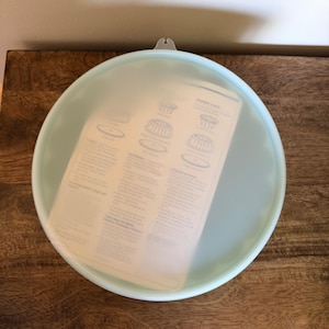 May include: A round, translucent, light blue silicone stretch lid with a small tab at the top edge. The lid is placed over a white instruction sheet that features diagrams and text detailing the use of various silicone food covers.