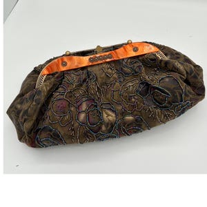 May include: A vintage-style clutch purse with a floral pattern in shades of brown, burgundy, and gold. The purse features beaded accents and an orange accent across the top. The clasp is gold-toned.