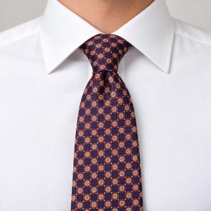 May include: A navy blue necktie featuring a repeating pattern of small circular motifs with red borders and white centers, worn with a crisp white collared dress shirt. The tie is tied in a neat knot at the center of the shirt collar.