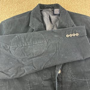 May include: A dark navy corduroy blazer with elbow patches and four buttons on the cuff. The jacket has a notched lapel and a single button closure. The fabric has a ribbed texture.