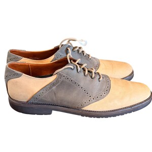 May include: A pair of two-tone oxford shoes featuring a tan suede body with grey suede overlays. The shoes include decorative brogue perforations along the seams, matching tan laces, a brown interior lining, and a dark brown rubber sole.