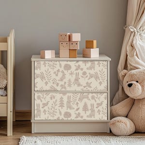 May include: A two-drawer dresser with a cream-colored finish and a pink woodland animal print design. The dresser is decorated with wooden blocks featuring smiley faces.