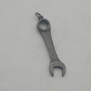May include: A small, silver-colored wrench pendant with a circular opening at one end and a U-shaped opening at the other. The pendant has a textured surface and a small loop for attaching to a chain or keychain.