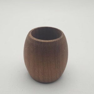 May include: A small, wooden, oval-shaped container with a dark brown finish. The container has a smooth exterior and a hollow interior. The wood grain is visible on the surface. The container is likely used for storage or display.