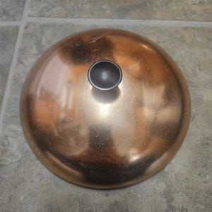 May include: A circular, dome-shaped copper lid with a polished metallic finish and a small, centered black knob handle. The surface of the lid shows light surface scratches and reflections, indicating a smooth, reflective metallic material.