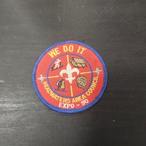 May include: A circular patch with a red background and blue border. The patch features the text "WE DO IT" at the top and "HEADWATERS AREA COUNCIL EXPO - 90" around the bottom. The center has a fleur-de-lis and various activity icons.