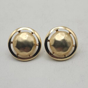 May include: A pair of gold-toned stud earrings. Each earring features a textured, circular center piece surrounded by a circular frame with small connecting bars. The earrings have a vintage or retro aesthetic.