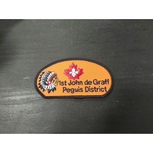 May include: An orange patch with black trim featuring a Native American headdress on the left and a red maple leaf with a fleur-de-lis in the center. The text "1st John de Graff Peguis District" is printed in black.
