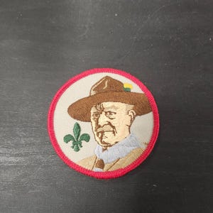 May include: Embroidered patch featuring a portrait of a man in a brown hat and uniform. The patch has a red border and a green fleur-de-lis symbol. The man's portrait is in the center of the patch.