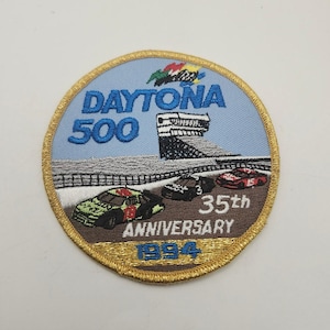 May include: A circular embroidered patch featuring a light blue background with the text DAYTONA 500 in blue. The design includes three race cars on a track, a grandstand, and the text 35th ANNIVERSARY 1994. The patch has a gold stitched border.