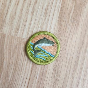 May include: Embroidered patch featuring a leaping fish design. The patch has a circular shape with a green border. The fish is depicted in shades of green and brown, with blue water details.
