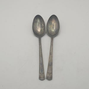 May include: Two vintage silver spoons with oval bowls and long, slender handles. The spoons have a tarnished appearance, suggesting age and use. The handles feature a decorative design near the base.