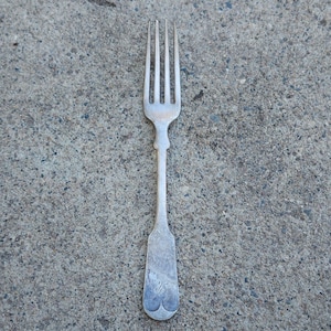 May include: A silver fork with four tines, a long handle, and a decorative design. The fork is resting on a gray, textured surface. The handle has a rounded end with a decorative design.