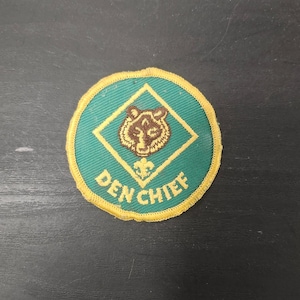 May include: A round patch with a green background and a gold border. The patch features a brown bear head inside a gold diamond, with a fleur-de-lis above it. The words "DEN CHIEF" are written in gold.