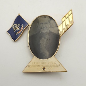 May include: A vintage pin featuring a black and white portrait of a smiling man in an oval frame. The pin includes a North Dakota flag, musical notes, and the text "Lawrence Welk North Dakota Roughrider 1969".