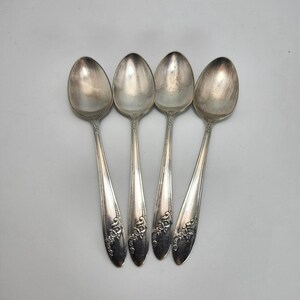 May include: Four silver spoons with ornate floral designs on the handles. The spoons are arranged in a row, with the bowls facing up. The spoons have a slightly tarnished appearance.