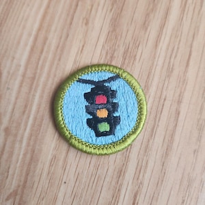 May include: A round embroidered patch featuring a traffic light design. The patch has a light blue background with a green border. The traffic light has red, yellow, and green lights. The patch is likely for a jacket or bag.