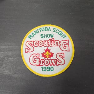 May include: A circular patch with a yellow border and white background. The patch features the text "Manitoba Scout Show" in green and "Scouting Grows 1990" in red. A red and yellow maple leaf is in the center.