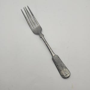 May include: A vintage silver fork with four tines. The fork has a long, slender handle and a rounded end. The metal has a slightly tarnished appearance, suggesting age and use. The fork is likely for eating.