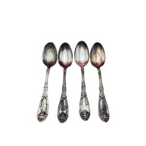 May include: Four antique silver spoons with ornate handles and oval bowls are arranged against a white background. The spoons show signs of age and use, with tarnishing on the silver. The handles feature detailed floral and vine patterns.