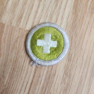 May include: Embroidered patch featuring a white cross on a lime green background, encircled by a white border. The patch is circular and has a textured appearance. The design is simple and clean, with a focus on the medical symbol.