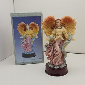 May include: A detailed angel figurine with outstretched wings, wearing a yellow top and a pink dress. The angel has brown hair and is standing on a dark brown base. A box with a similar image is next to the figurine.