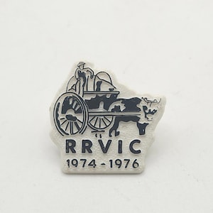 May include: A vintage pin featuring a black and white illustration of a farmer with a cart pulled by a cow. The pin has the text "RRVIC 1974-1976" in black lettering on a white background. The pin is a collectible item.